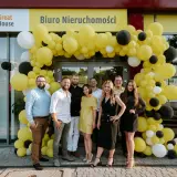 Grupa ludzi pozuje przed biurem nieruchomości ozdobionym żółtymi i czarnymi balonami.