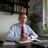 Mężczyzna w koszuli i krawacie siedzi przy biurku, pisząc w notatniku. Na biurku laptop, kalkulator i myszka.