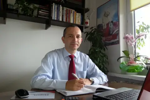Mężczyzna w koszuli i krawacie siedzi przy biurku, pisząc w notatniku. Na biurku laptop, kalkulator i myszka.
