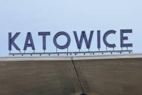 Napis "KATOWICE" z niebieskimi neonami na tle jasnego nieba, nad betonową konstrukcją.