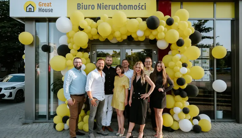 Grupa ludzi pozuje przed biurem nieruchomości ozdobionym żółtymi i czarnymi balonami.
