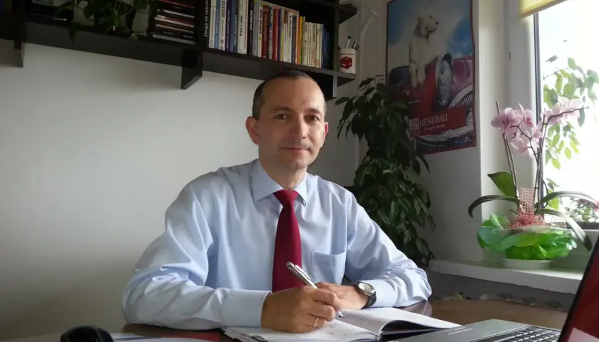 Mężczyzna w koszuli i krawacie siedzi przy biurku, pisząc w notatniku. Na biurku laptop, kalkulator i myszka.