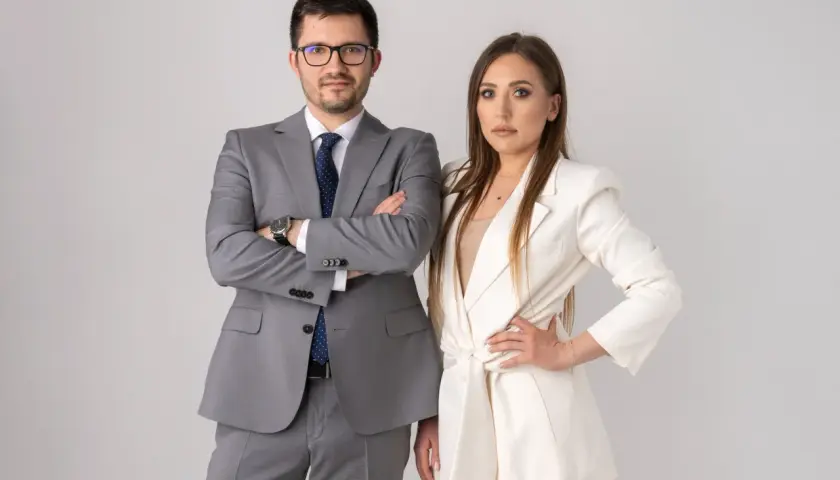 Para w garniturach, mężczyzna z rękami skrzyżowanymi na piersi, kobieta z ręką na biodrze.