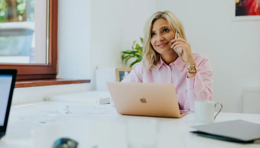 Kobieta w różowej koszuli rozmawia przez telefon przy biurku z laptopem.