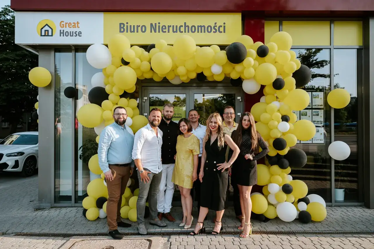 Grupa ludzi pozuje przed biurem nieruchomości ozdobionym żółtymi, czarnymi i białymi balonami.